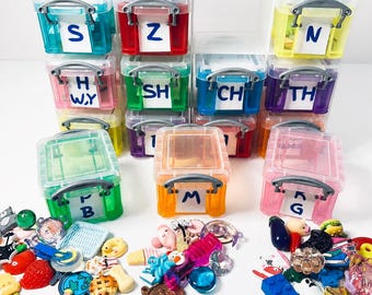 May include: A collection of small, colorful, translucent plastic storage containers with silver handles, each labeled with white paper tags featuring black letters or letter combinations. The containers are filled with various miniature charms.