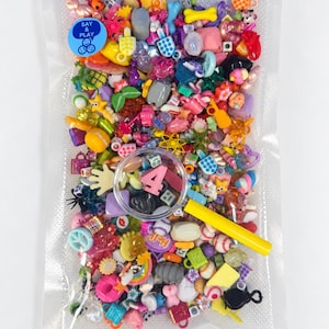 May include: A clear plastic vacuum-sealed bag filled with a variety of colourful miniature plastic charms and trinkets in diverse shapes. A yellow-handled magnifying glass rests on top, highlighting a pink number four charm inside the bag.