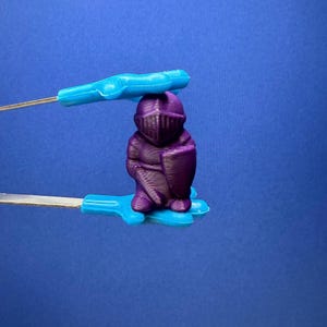 May include: A small, purple knight figurine with a shield and helmet is held by blue tweezers against a blue background. The knight is a solid color and has a detailed design. The tweezers are made of metal and plastic.