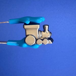 May include: A small, silver-colored toy train held by blue plastic tongs against a blue background. The train has a classic design with circular wheels and a smokestack.