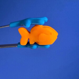 May include: A bright orange fish-shaped object held by a pair of metal tongs with blue silicone tips. The fish has a smooth, matte surface and a distinct fin and tail structure. The tongs are positioned horizontally, gripping the orange fish firmly.