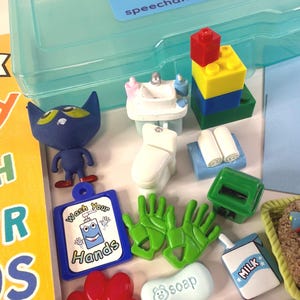May include: A collection of colorful toy figures and accessories, including a blue cat, a miniature sink, a toilet, a stack of blocks, green gloves, and a sign that says "Wash Your Hands". A blue plastic container is in the background.