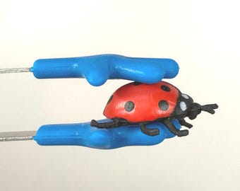 Può includere: Una statuetta di coccinella rossa e nera con punti neri e antenne, tenuta tra due manici di plastica blu. La coccinella è il punto focale, con i manici blu che forniscono uno sfondo contrastante.