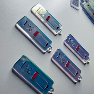 May include: Six iridescent keychains shaped like phone battery icons, each displaying "1%" and a red battery bar. The keychains are arranged in a semi-circle on a white surface.