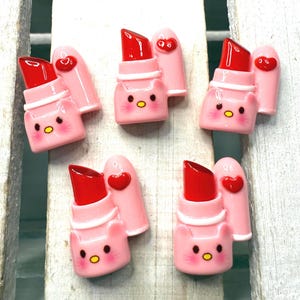 May include: Five pink and red lipstick charm pendants with a cartoon cat face. The lipstick is red and the tube is pink with a white stripe. The cat face has pink cheeks and a yellow nose.