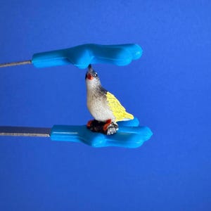 May include: A miniature bird figurine with yellow, black, and white plumage perched on a small, dark base. The bird is held by a blue tool with a handle. The background is a solid blue.