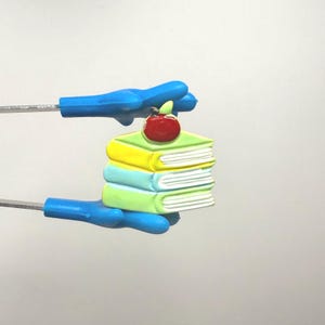 May include: A stack of three colorful books with a red apple on top. The books are yellow, blue, and green. The apple is red with a green leaf. The books are held by blue handles.
