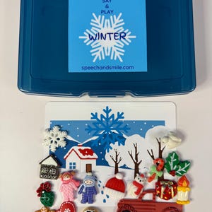 Puede incluir: Una caja de almacenamiento de plástico azul con una etiqueta que dice "SAY & PLAY WINTER speechandsmile.com". La caja está abierta, revelando elementos decorativos con temática invernal, incluyendo un camión rojo, una casa y figuras.