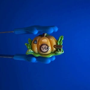 Könnte beinhalten: Miniatur-Kürbis-Haus-Figur mit gelbem Körper, grünen Blättern und einer kleinen Tür. Das Haus hat ein rundes Fenster und ein Schild mit der Aufschrift "HAPPY". Die Figur wird von blauen Werkzeugen vor blauem Hintergrund gehalten.