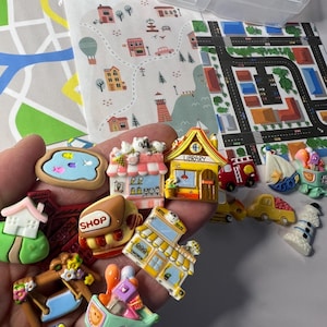 Puede incluir: Una colección de coloridos edificios de juguete en miniatura, vehículos y otros objetos, que incluyen una biblioteca, una tienda, una cafetería, un taxi, un carrito de compras y un faro. Los juguetes están hechos de plástico y tienen una variedad de diseños.