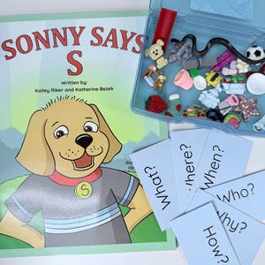 以下が含まれることがあります： 「Sonny Says S」というタイトルの子供向けの本で、漫画の子犬のイラストが描かれています。本の隣には、疑問詞が書かれたフラッシュカードと、サッカーボールやヘビのおもちゃなど、小さなおもちゃや装身具で満たされた透明なプラスチック容器があります。