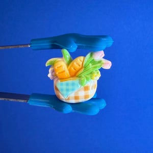May include: A miniature basket with a gingham pattern, filled with carrots, bread, and flowers. The basket is held by blue tools against a blue background. The basket is a decorative item.