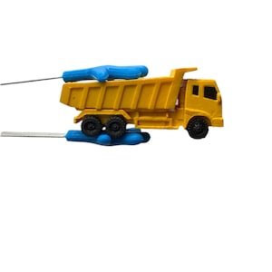 Puede incluir: Un camión volquete de juguete amarillo con ruedas negras y un asa azul. El camión tiene un diseño detallado, incluyendo una cabina y una caja volquete. El juguete está sobre un fondo blanco.