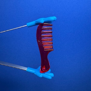May include: A translucent red comb with a curved handle and evenly spaced teeth. The comb is held by two blue plastic hands attached to silver wires, set against a solid blue background. The comb is a hair care accessory.