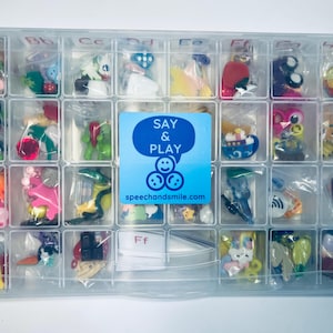 Könnte beinhalten: Eine transparente Plastik-Aufbewahrungsbox mit 24 Fächern, gefüllt mit kleinen bunten Spielzeugen. Jedes Fach hat einen durchsichtigen Plastikbeutel mit einem Spielzeug darin. Die Box hat ein blaues Etikett mit der Aufschrift "SAY & PLAY" und "speechandsmile.com".