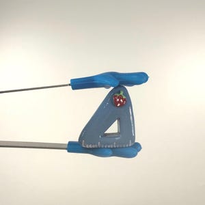 Può includere: Uno strumento a forma di triangolo blu chiaro con un design a fragola rossa. Lo strumento ha un'apertura triangolare ed è tenuto da manici blu. Lo strumento viene utilizzato per tagliare o modellare materiali.