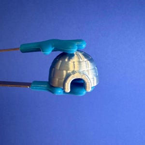 May include: A silver igloo model with a curved entrance is held by blue plastic tongs against a blue background. The igloo has a domed roof with rectangular block details.