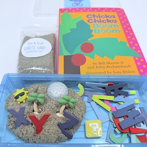 Puede incluir: Un contenedor de plástico azul lleno de arena cinética y fichas de letras del alfabeto de colores. El contenedor también incluye un pequeño sol de plástico, dos palmeras y un huevo plateado. El libro "Chicka Chicka Boom Boom" de Bill Martin Jr. y John Archambault, ilustrado por Lois Ehlert, es visible en el fondo.