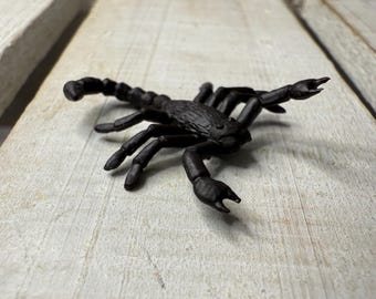May include: A black plastic scorpion toy with detailed legs, pincers, and a curved tail. The scorpion is positioned on a weathered, light-colored wooden surface. The toy is a realistic representation of the arachnid.
