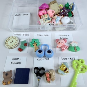 May include: A clear plastic storage container filled with assorted miniature plastic charms and buttons. Below the container are white cards with word pairs like clock-sock, tree-see, mouse-house, bear-square, cut-nut, and bee-key, each paired with matching charms.