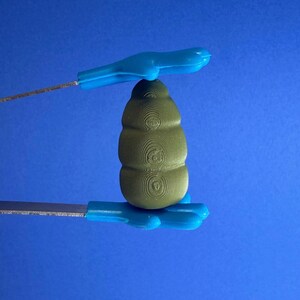 May include: A green, textured object with a stacked design is held by two blue tools against a blue background. The tools have a handle and a thin, metallic blade. The object appears to be a unique tool or component.
