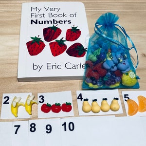 May include: An educational set for children, including a book titled "My Very First Book of Numbers" by Eric Carle, and numbered cards with fruit-shaped decorations. A blue mesh bag contains more fruit-themed decorations. Numbers 1 to 10 are displayed.