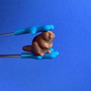 May include: A brown beaver figurine held by blue tongs against a blue background. The beaver has a detailed tail and is positioned between the tongs. The tongs have a cloud-like shape at the top and bottom.