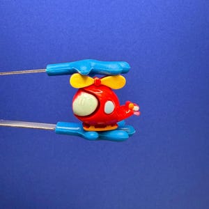 May include: A red toy helicopter with yellow blades and a light green window. The helicopter is held by two blue plastic handles connected by thin, silver wires, against a blue background.