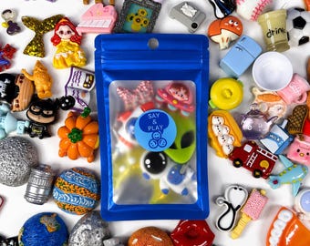 May include: A blue resealable pouch with a clear window containing various small plastic charms, featuring a circular sticker with the text Say & Play. The pouch is surrounded by a collection of colorful miniature charms including animals, food, and space-themed items.