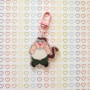 May include: A novelty keychain with a cartoon character and a pink clasp. The character wears glasses, a white shirt, green trousers, and has a brown tail. The keychain is set against a white background with colourful heart shapes.