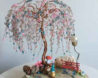 May include: A whimsical wire tree sculpture with pink and blue beads, a small gnome figurine, a red bench, and a mushroom under a weeping willow tree. The sculpture is on a grey rock base.