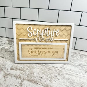 May include: A rectangular wooden sign with a white frame. The sign features the words "Scripture of the week" in a cursive font. Below, it reads "Forgive one another just as God forgave you" from Ephesians 4:32.
