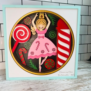 Puede incluir: Una obra de arte de madera en capas que presenta a una bailarina con un vestido rosa con detalles de bastones de caramelo, rodeada de caramelos rojos y verdes y un poste a rayas rojas y blancas. La obra está enmarcada en un marco azul claro, con el texto "Elianore as Clara The Nutcracker - 2023".