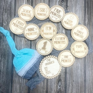 May include: Wooden baby milestone discs with months one through eleven, plus "I've Arrived" disc. A blue, white, and gray knit baby hat rests on a gray blanket. The discs have a checkered border and skateboard graphics.