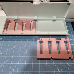 Pode incluir: Um porta-ferramentas de pl&aacute;stico de cor verde claro e rosa com cinco ranhuras para guardar ferramentas. O porta-ferramentas est&aacute; aberto e as ranhuras est&atilde;o vazias. O porta-ferramentas est&aacute; sobre um tapete de corte azul.