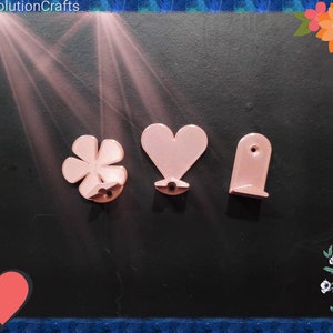 May include: Three pink plastic wall hooks in the shapes of a flower, a heart, and a rectangle. The hooks are on a black background.