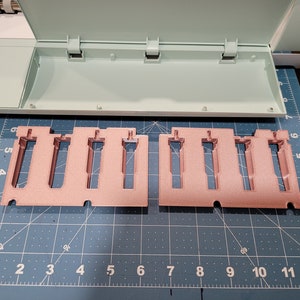 Pode incluir: Dois porta-ferramentas de pl&aacute;stico rosa com ranhuras para segurar ferramentas. Os suportes est&atilde;o sobre uma superf&iacute;cie de grade azul e branca.