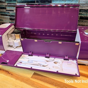 May include: A purple and white crafting machine with an open lid, revealing a compartment with crafting tools. The machine has a control dial and a front panel that opens to display tools. The text "Tools Not Included" is visible.