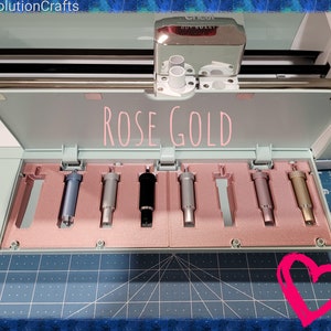 Pode incluir: Ferramentas de corte Cricut cor ouro rosa em uma caixa de armazenamento. As ferramentas est&atilde;o dispostas em uma fila e todas s&atilde;o do mesmo tamanho. A caixa est&aacute; aberta e as ferramentas s&atilde;o vis&iacute;veis. A caixa &eacute; feita de pl&aacute;stico azul claro e tem um interior rosa.