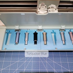 Pode incluir: Close-up de um conjunto de sete l&acirc;minas de corte de diferentes cores para uma m&aacute;quina de corte artesanal. As l&acirc;minas est&atilde;o dispostas em uma fila em um suporte de espuma azul. O texto "Turquoise Blue" &eacute; vis&iacute;vel abaixo das l&acirc;minas.