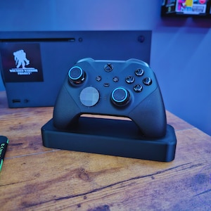 May include: A black Xbox controller with blue accents and a textured grip. The controller is sitting on a black charging stand. The controller has a  "Wounded Warrior Project" sticker on the front.