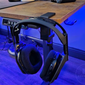 May include: Two black headphones with padded earcups are hanging from a desk mounted headset holder. The desk is made of wood and has a blue light shining on it.