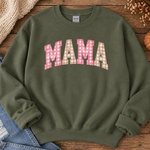 Pode incluir: Uma camisola de gola redonda verde-azeitona com a palavra MAMA numa fonte arqueada de estilo universitário. As letras têm um padrão de xadrez vichy rosa e branco que alterna com um padrão de xadrez vichy bege e branco.
