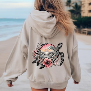 Puede incluir: Una sudadera con capucha de color beige que presenta un gráfico en la parte posterior. El diseño muestra la silueta de una tortuga marina rellena con una ola del océano, un atardecer y una flor de hibisco rosa, enmarcada por hojas estilizadas.
