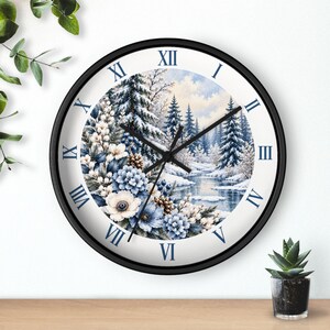 May include: A round wall clock with a black frame and white face featuring Roman numerals in dark blue. The center displays a winter landscape with snow-covered pine trees, a frozen stream, and a floral arrangement of blue and white flowers.