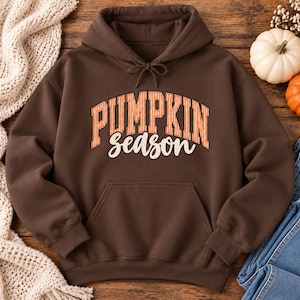 May include: A dark brown hooded sweatshirt featuring a front kangaroo pocket and a drawstring hood. The chest displays the text PUMPKIN in an orange striped collegiate font, with the word season written below in a cream-colored cursive script.