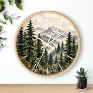 May include: A round wall clock with a light wood frame featuring a watercolor-style illustration of a dense pine forest and snow-capped mountains in the background. The clock face has simple white hour markers and white clock hands.