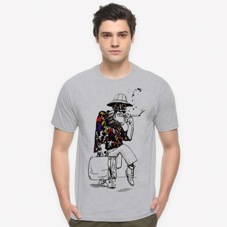 May include: A light grey t-shirt with a black and white illustration of a smoking figure wearing a hat and patterned shirt. The figure is seated and holding a suitcase. The shirt has a crew neck.