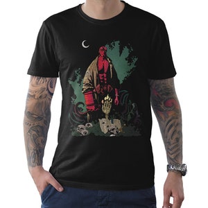 Puede incluir: Camiseta negra con un diseño de Hellboy. El diseño incluye una figura roja de Hellboy, calaveras y una mano brillante. La obra de arte está sobre un fondo verde oscuro y negro con una luna creciente.