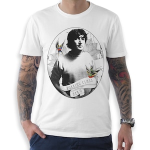 May include: White t-shirt featuring a black and white portrait inside an oval frame. The portrait is of a person with dark hair. The frame includes floral elements and two swallows. A banner below the portrait reads "WORKING CLASS HERO".